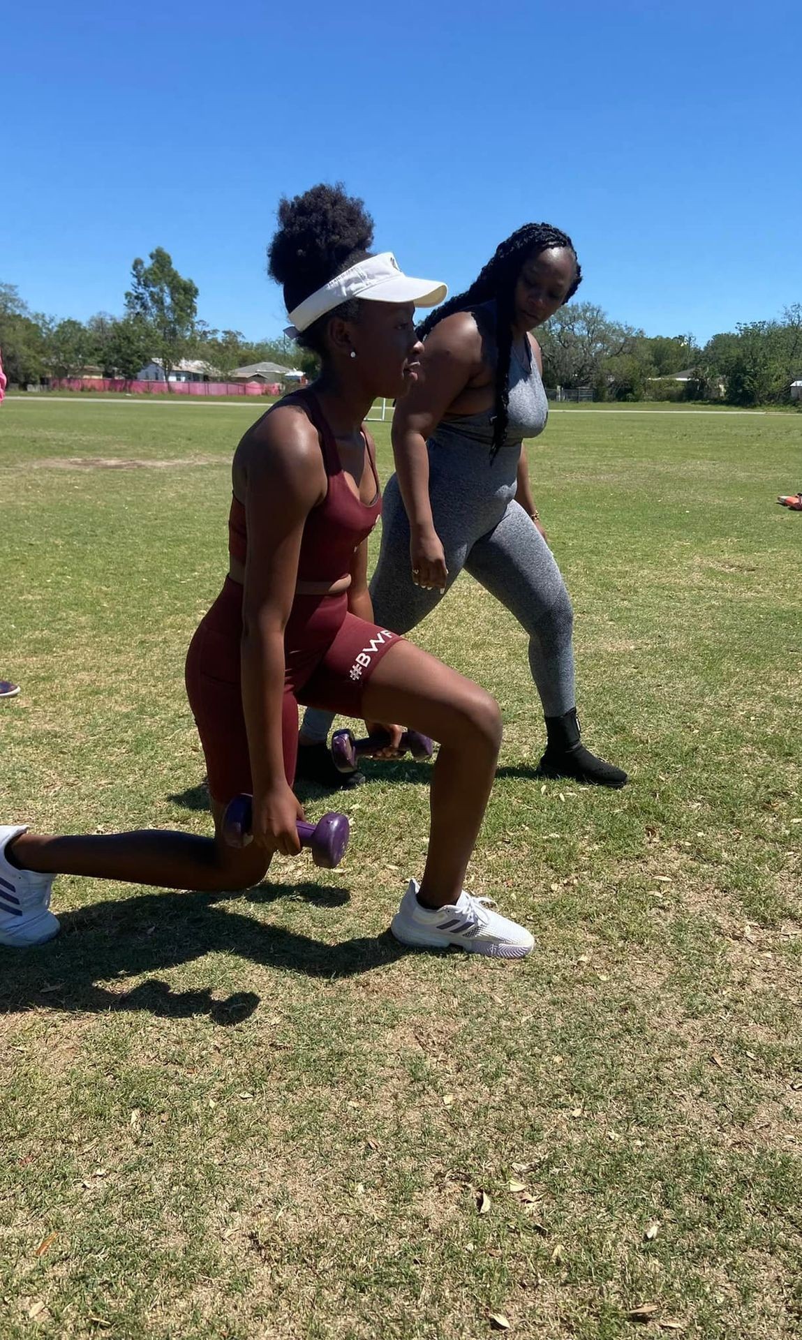 Two women exercising outdoors, one performing a lunge with dumbbells on a grassy field.